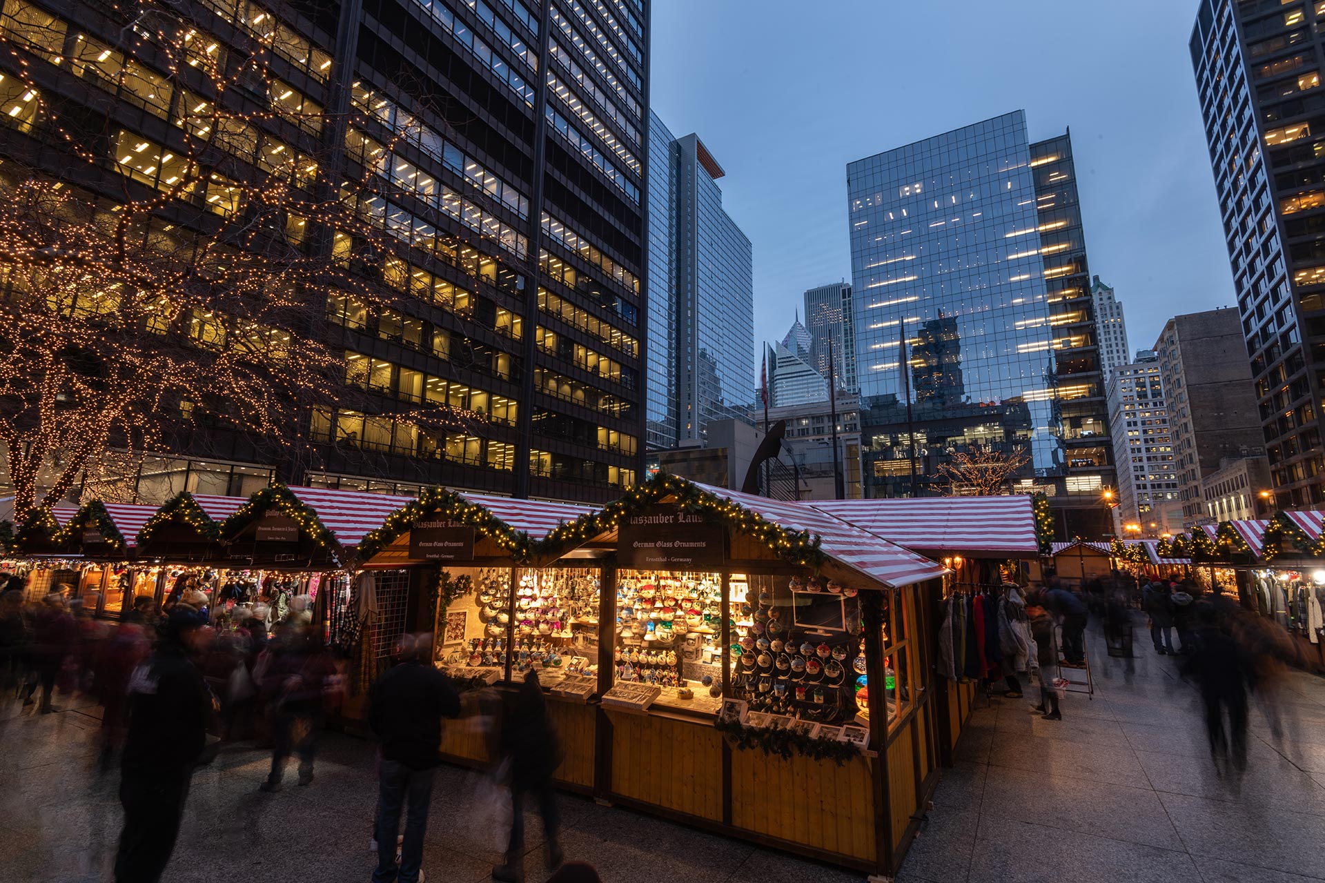  A bustling holiday Chriskindlmarket market at dusk in downtown Chicago, featuring festive wooden stalls adorned with twinkling lights, surrounded by tall skyscrapers.