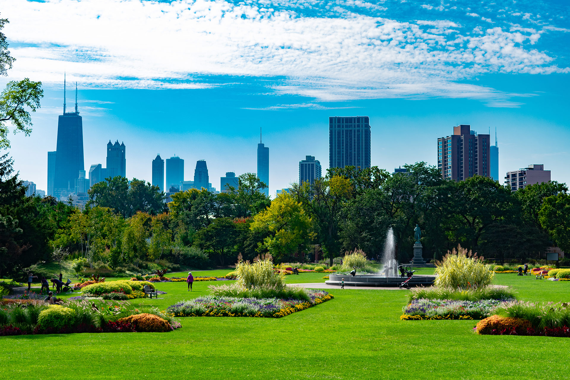 Scenic view of a vibrant park with lush greenery, colorful flower beds, a fountain, and Chicago's skyline in the background on a sunny day.