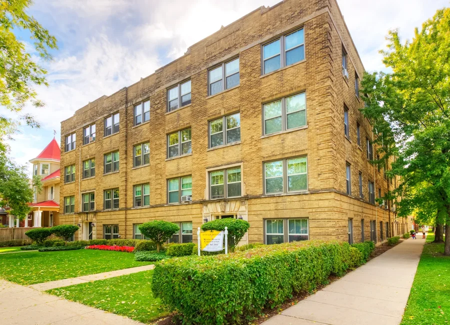 Exterior view of a vintage brick apartment building with trimmed hedges and a sidewalk in front, taken on a bright, partly cloudy day.
